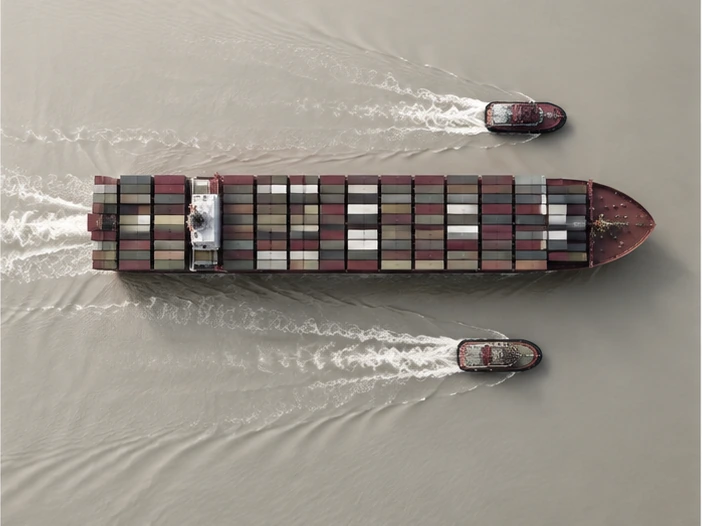 A container ship and two tugboats on a light grey sea, seen from above