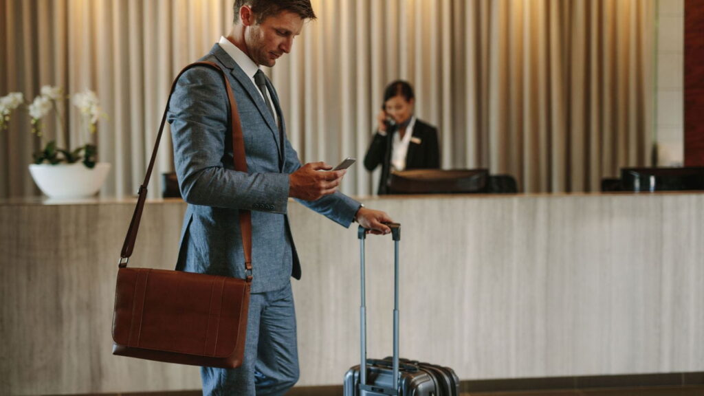 Homme d’affaires en costume consultant son téléphone à la réception d’un hôtel avec une valise.