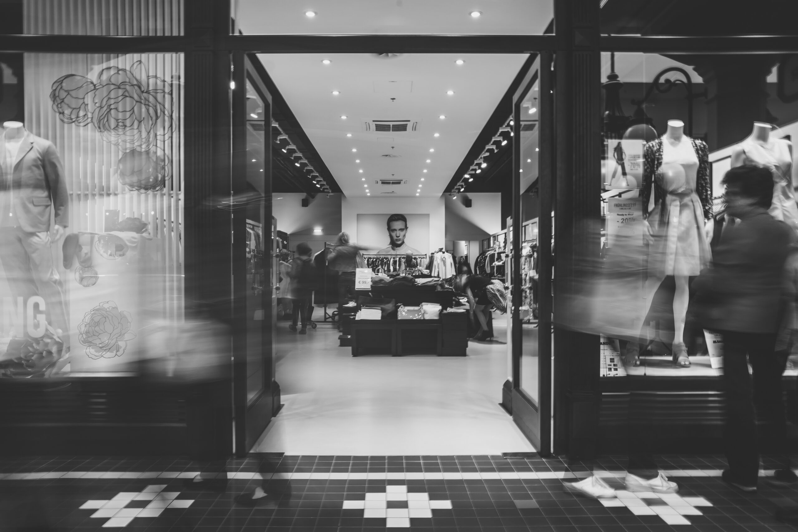 Entrée d’une boutique de vêtements en noir et blanc, avec des mannequins en vitrine et des clients en mouvement.