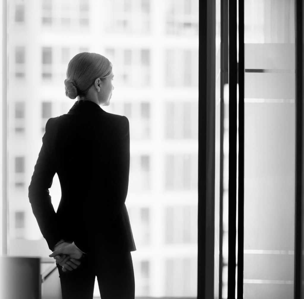 Femme en tailleur noir observant la vue depuis une fenêtre dans un bureau lumineux.