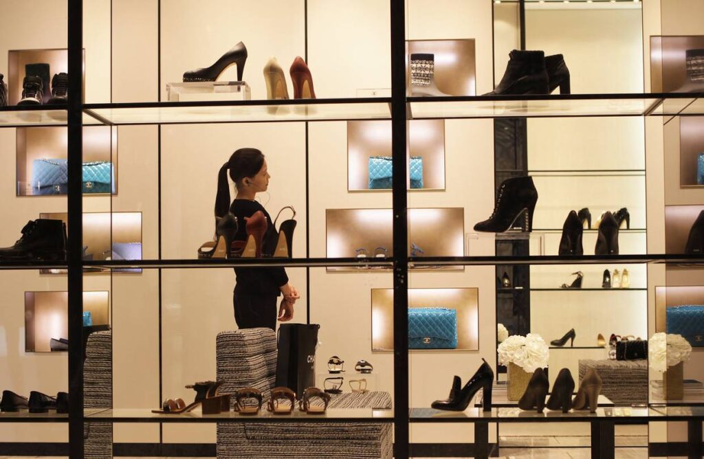 Vitrine d’une boutique de luxe présentant des chaussures, sacs à main et accessoires, avec une vendeuse à l’intérieur.