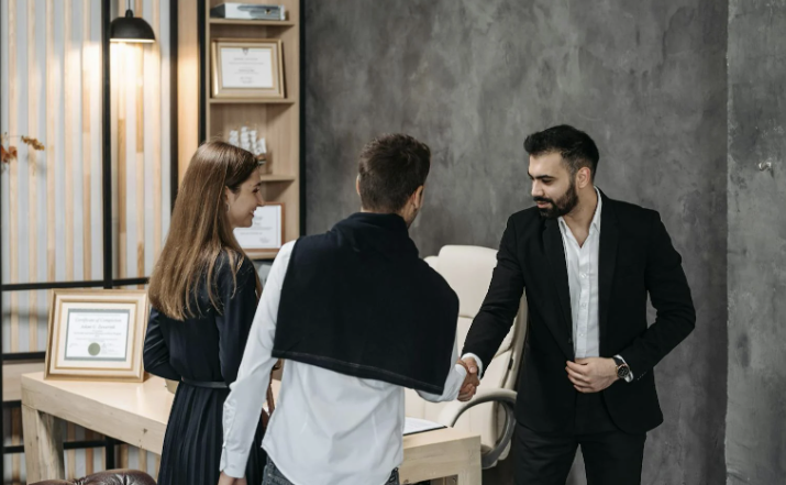 Homme en costume serrant la main d’un client dans un bureau, en présence d’une femme.
