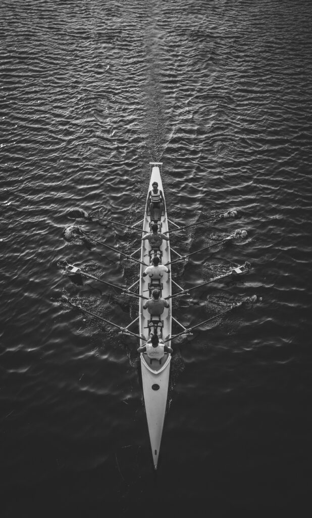 Vue aérienne en noir et blanc d’un bateau d’aviron avec cinq rameurs avançant sur l’eau.